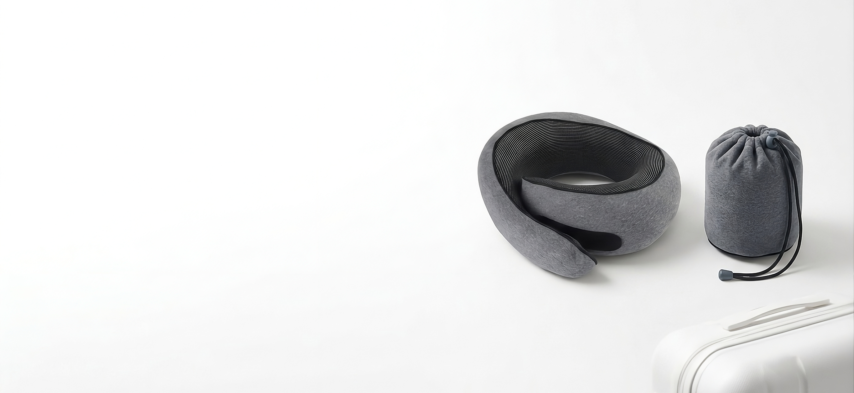 A gray neck pillow with a black inner lining is shown next to its matching gray drawstring storage pouch on a clean white background. A white suitcase is partially visible in the bottom right corner.